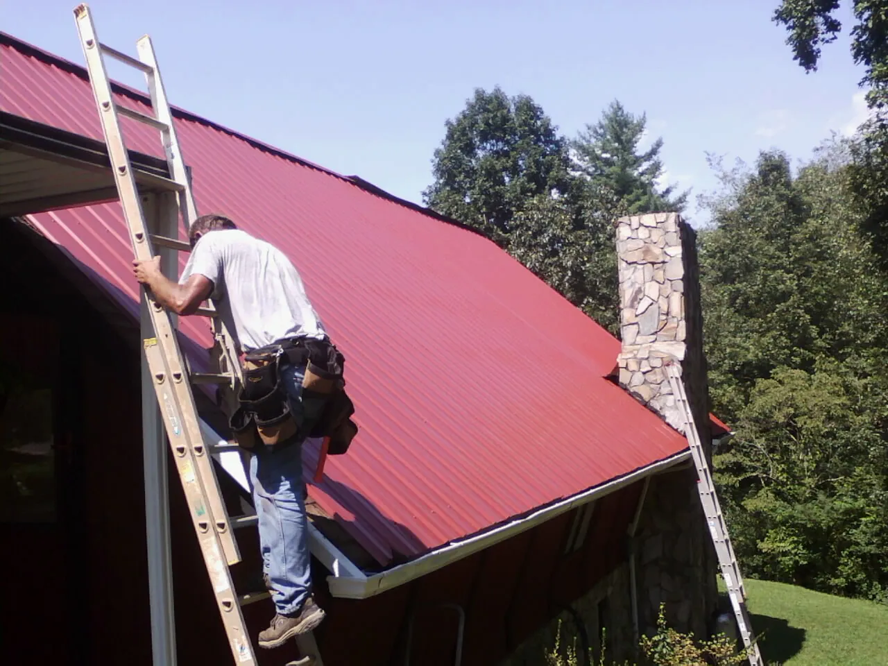 Roofer on a ladder beside a red metal roof and stone chimney — Soffit & Fascia Repair by Roofing Brothers in Beasley